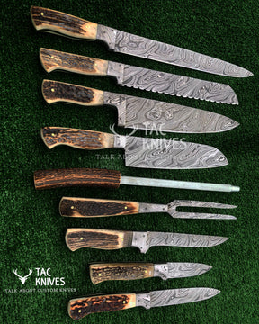 A set of custom knives and a carving fork arranged diagonally on a green textured background. The knives feature patterned Damascus steel blades and handles made from natural deer horns with dark brown and tan sections.