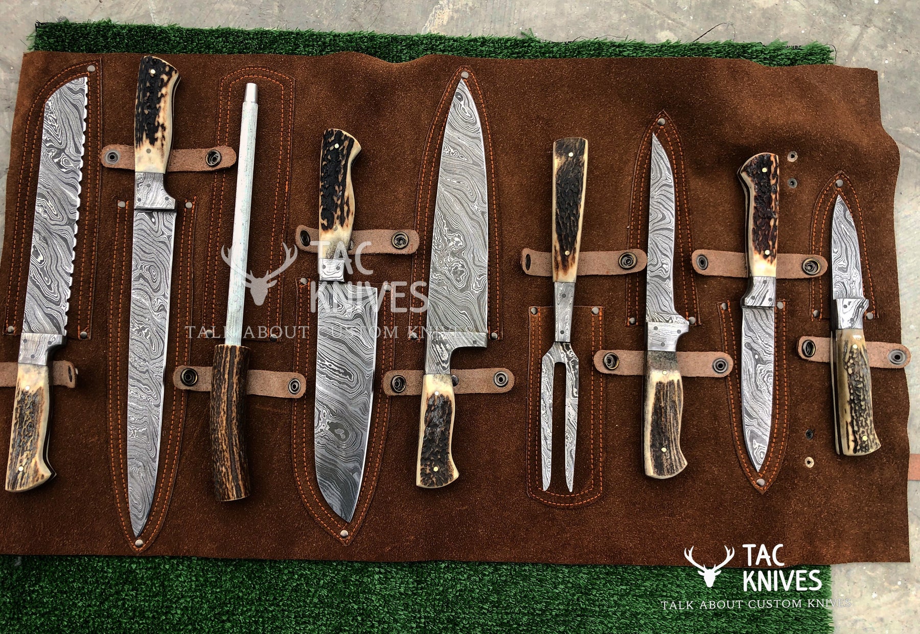 A set of seven custom knives with Damascus steel blades and butchered cowhide handles, displayed on a brown suede knife roll over green artificial turf. The brand 'Tac Knives' is visible as a watermark and logo.