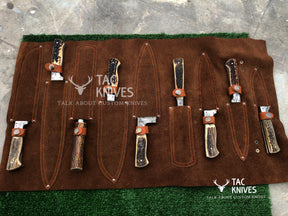A brown suede knife roll holding multiple custom knives with dark wood and bone handles, displayed on a green artificial turf mat over a gray concrete floor. The Tac Knives logo is visible on the roll.