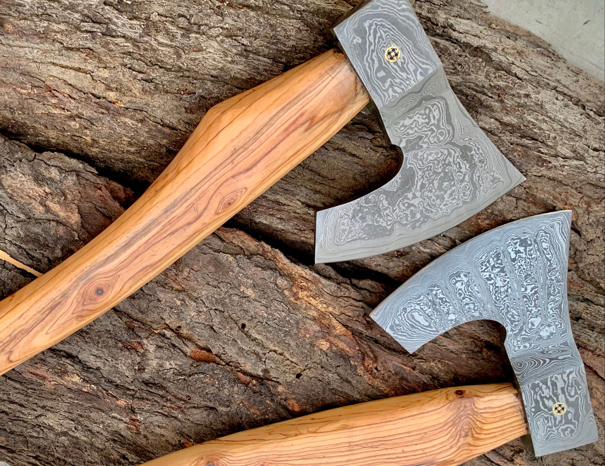 Two hand-crafted bearded axes with Damascus steel heads and natural wood handles, displayed diagonally on a textured tree trunk background.