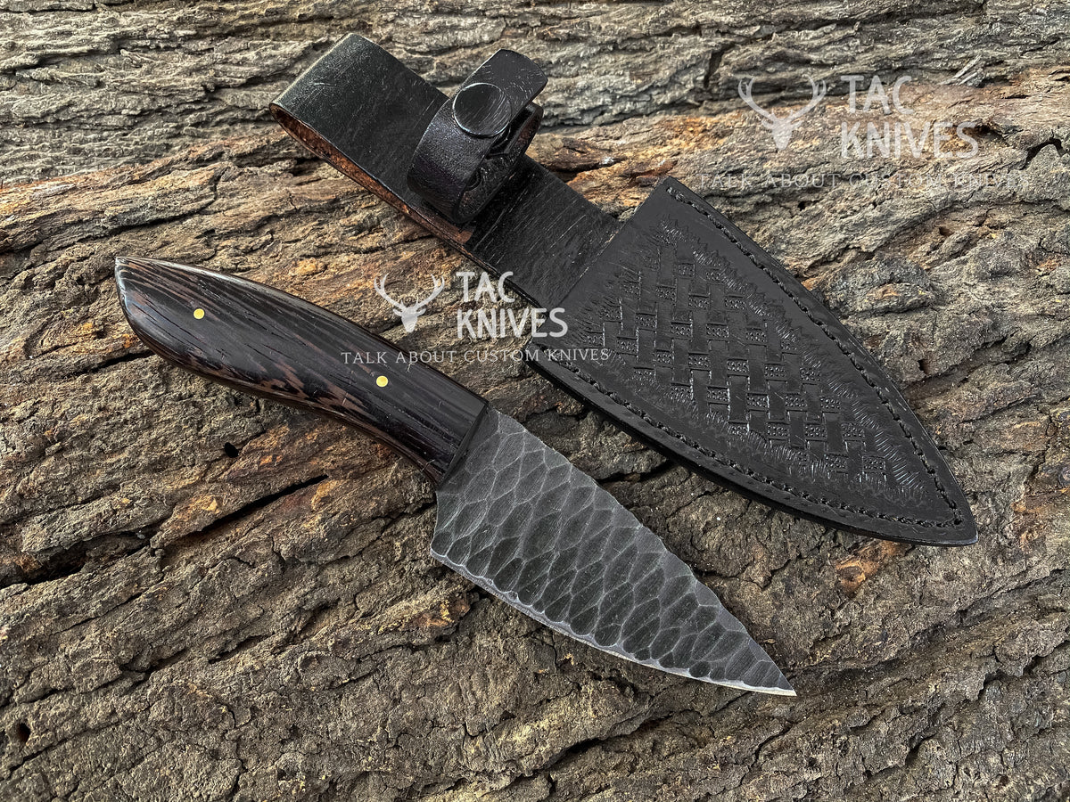 A fixed-blade knife with a dark wood handle and hammered steel blade, displayed next to its black woven leather sheath on a textured tree bark background. The Tac Knives logo is visible on the image.