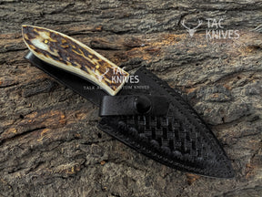 A custom handcrafted pocket knife with a natural bone handle featuring dark brown and cream marbling, shown in its black woven leather sheath on a textured tree bark background. The Tac Knives logo is visible on the handle and as an overlay.