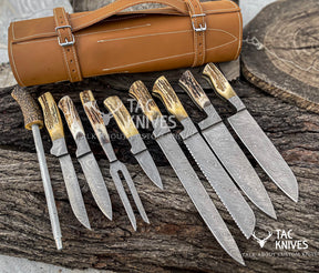 A set of eight custom hunting knives with patterned Damascus steel blades and antler-style handles, displayed alongside a tan leather knife roll on a rustic wood surface.