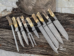 A set of custom handcrafted knives with patterned Damascus steel blades and natural antler handles, displayed on a rustic log slice with a sharpening steel.