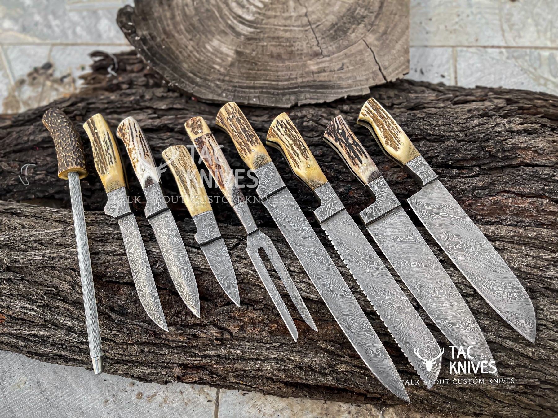 A set of nine custom knives and a sharpening steel arranged on a rustic log. The knives feature patterned Damascus steel blades and natural antler handles with golden-brown tips.