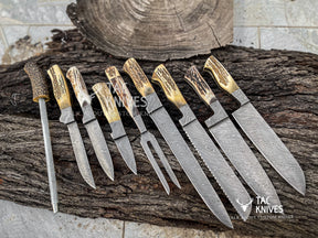 A set of nine custom knives and a sharpening steel arranged on a rustic log. The knives feature patterned Damascus steel blades and natural antler handles with golden-brown tips.