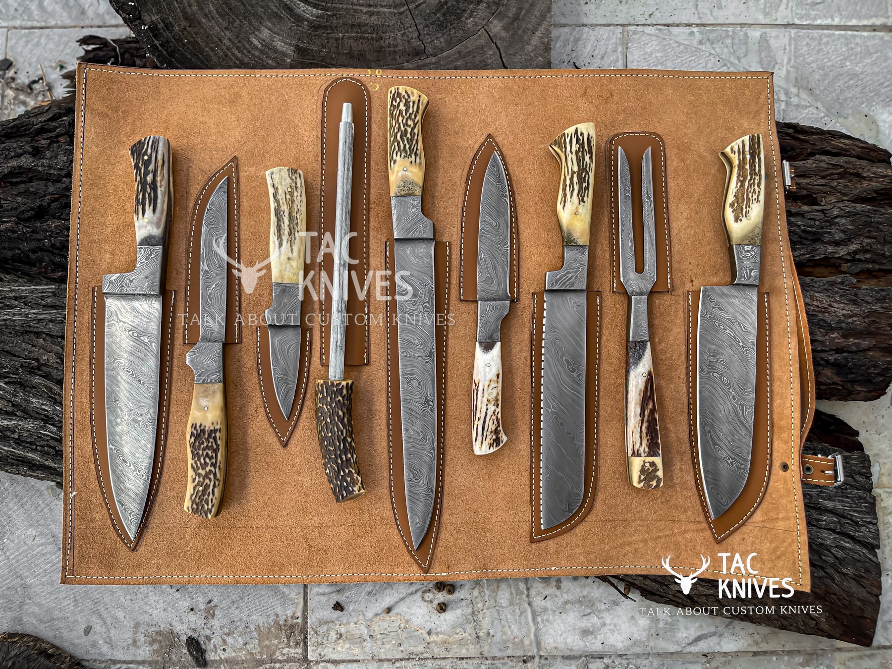 A set of nine custom hunting and butchery knives with patterned steel blades and natural antler or bone handles, displayed on a tan leather roll-up organizer. The knives are arranged on a rustic background of dark wood logs and gray stone tiles.