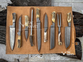 A set of nine custom hunting and butchery knives with patterned steel blades and natural antler or bone handles, displayed on a tan leather roll-up organizer. The knives are arranged on a rustic background of dark wood logs and gray stone tiles.