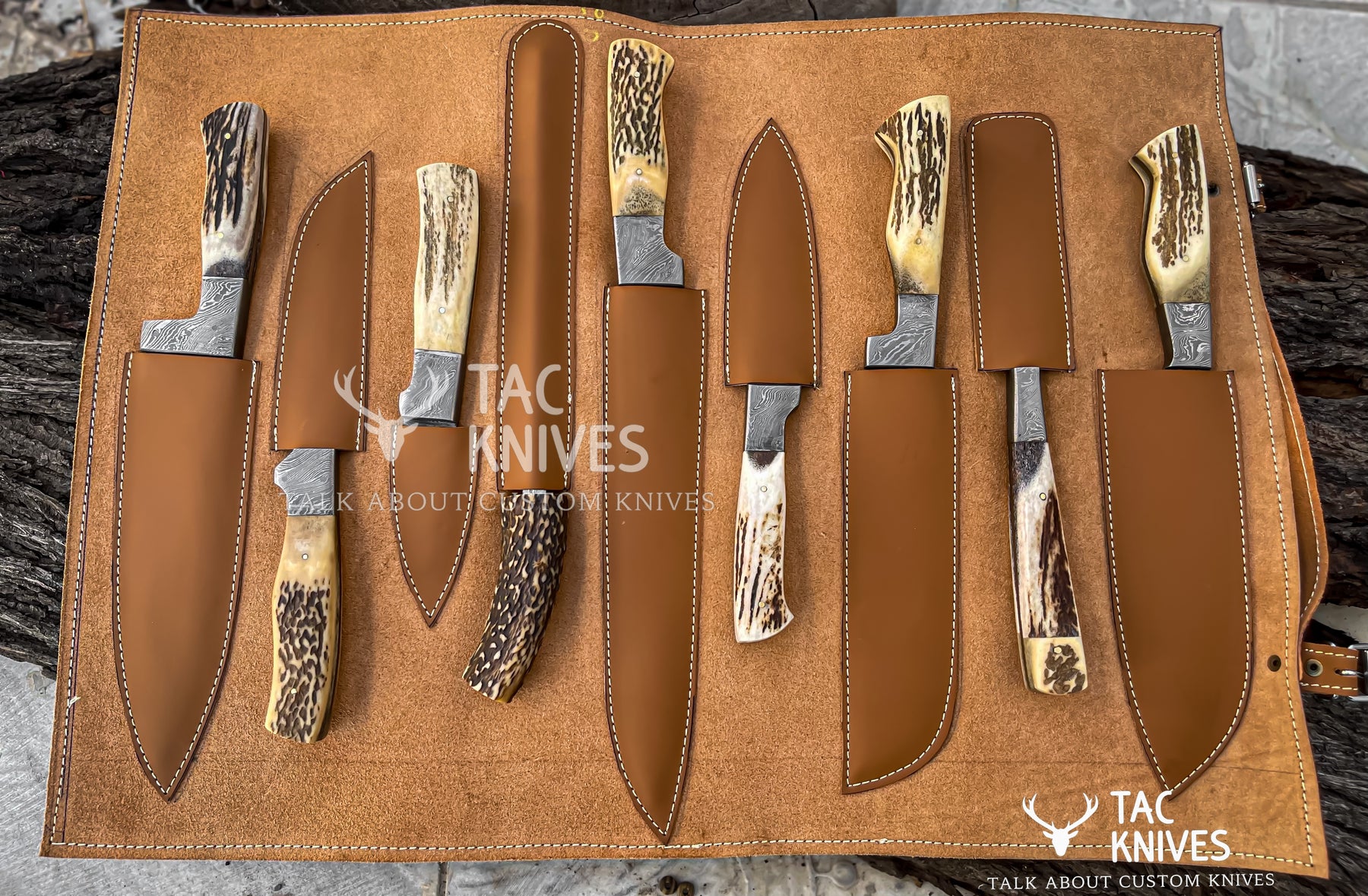 A set of custom knives with antler handles and steel blades, displayed in a brown leather roll-up case on a rustic wood background. The brand 'Tac Knives' is visible as watermarks and logos.
